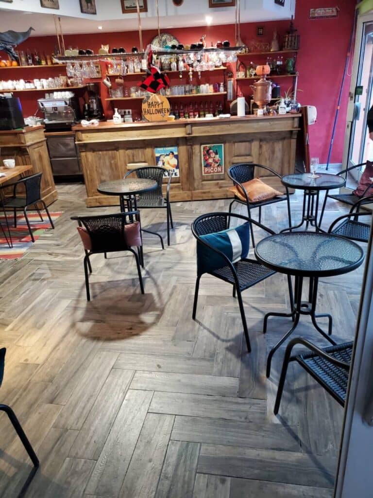 Bar-café ambiance Halloween : Décoration vintage et cosy Intérieur bar-café ambiance Halloween. Comptoir bois, étagères. Signe "Happy Halloween", affiche Picon. Tables rondes noires, chaises.