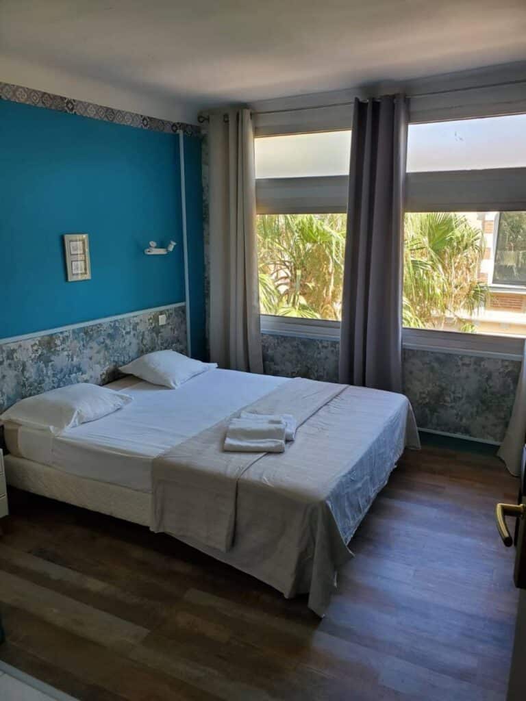 Hôtel: Chambre bleue moderne, lit double, vue palmiers Chambre d'hôtel lumineuse avec lit double, murs bleus, et grande fenêtre donnant sur des palmiers. Parquet en bois et rideaux gris.