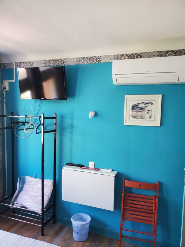 Chambre d'hôtel lumineuse : TV, Climatisation, Rangement Chambre moderne aux murs bleu turquoise avec TV, climatisation, étagère à vêtements et table murale rabattable. Chaise rouge.